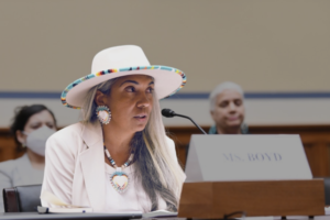 Kara Boyd at the congressional subcommittee on environment's hearing regarding regenerative agriculture on July 19th, 2022. She is wearing all white with beaded jewelry. She is speaking into a microphone, in front of a blurred, darker background containing two people.