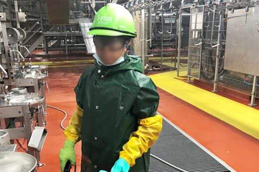 A child photographed by the Department of Labor during its investigation of Packers Sanitation Services Inc. (PSSI). The child is cleaning a slaughterhouse. The child is wearing a green hard hat that says PSSI, a dark green waterproof jacket, yellow arm sleeves, and gloves. The child's face has been blurred.